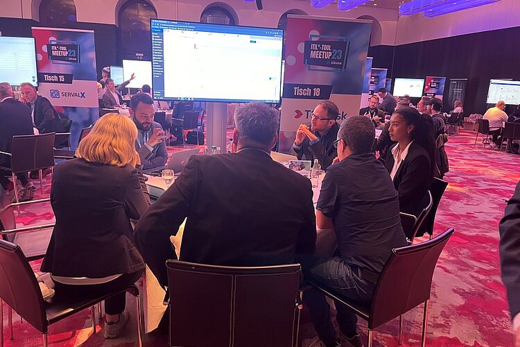 Participants sit at a round table during an ITIL Tool Meetup session, discussing and watching a presentation on a large screen.