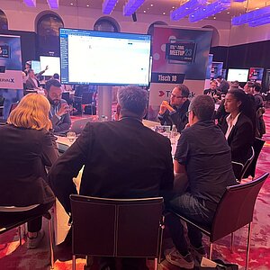 Participants sit at a round table during an ITIL Tool Meetup session, discussing and watching a presentation on a large screen.