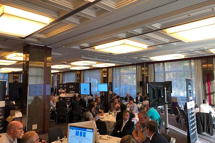 Participants sit at a round table during a tool meetup session, discuss and watch a presentation on a large screen.