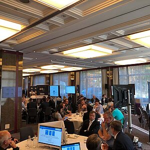 Participants sit at a round table during a tool meetup session, discuss and watch a presentation on a large screen.