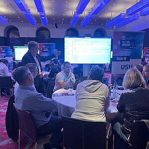 Participants sit at a round table during a tool meetup session, discuss and watch a presentation on a large screen.
