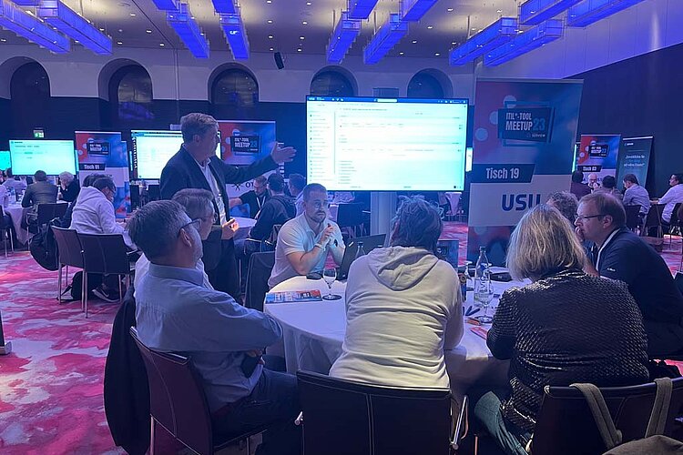 Participants sit at a round table during a tool meetup session, discuss and watch a presentation on a large screen.