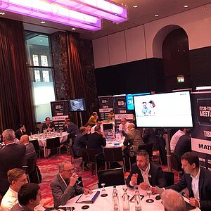 Participants in the ITSM tool MeetUp sit at round tables in a modern conference room with large screens and LED ceiling lighting.