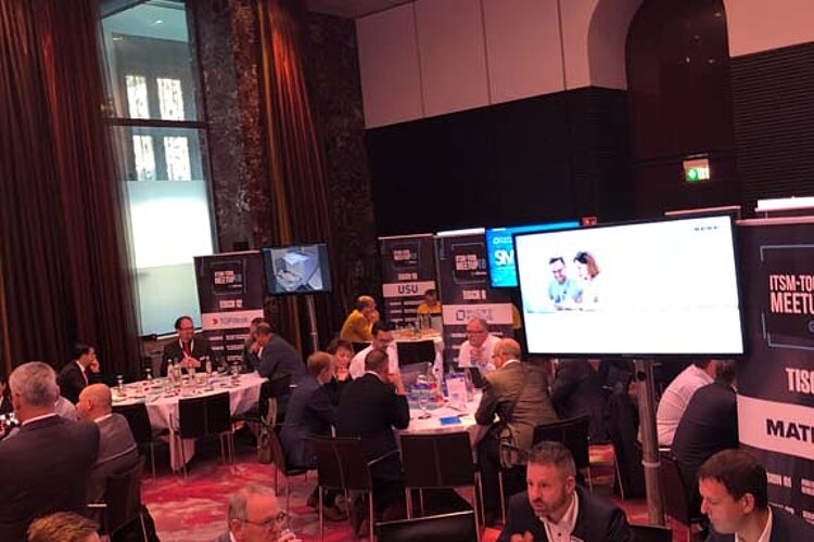 Participants in the ITSM tool MeetUp sit at round tables in a modern conference room with large screens and LED ceiling lighting.