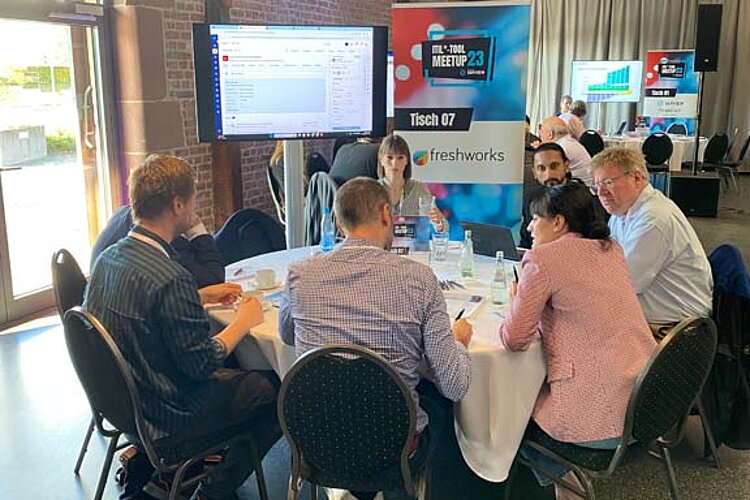 Participants sit at a round table during a tool meetup session, discuss and watch a presentation on a large screen.