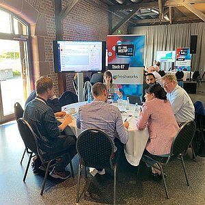 Participants sit at a round table during a tool meetup session, discuss and watch a presentation on a large screen.