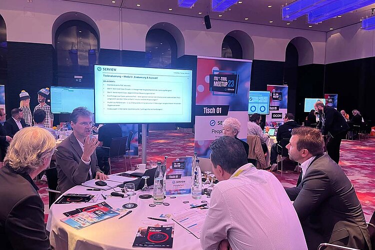 Participants at SERVIEW TOOL MEETUP23 discuss IT tool evaluation at a round table while a presentation is shown on a large screen.