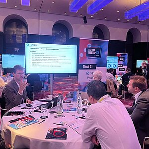 Participants at SERVIEW TOOL MEETUP23 discuss IT tool evaluation at a round table while a presentation is shown on a large screen.