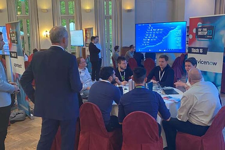 Participants sit at a round table during a tool meetup session, discuss and watch a presentation on a large screen.