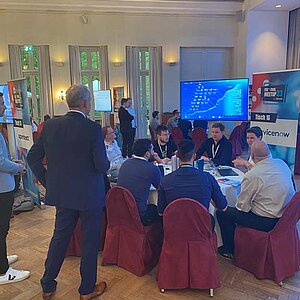 Participants sit at a round table during a tool meetup session, discuss and watch a presentation on a large screen.