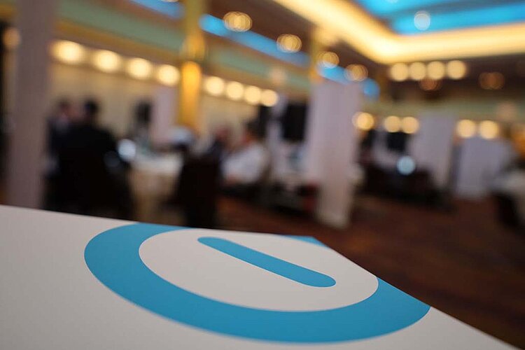 Close-up of the SERVIEW logo (blue circle with blue bar in the middle) on a white stand with a blurred background of a conference hall at the SERVIEW TOOL MEETUP.