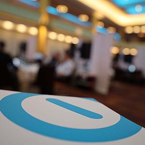 Close-up of the SERVIEW logo (blue circle with blue bar in the middle) on a white stand with a blurred background of a conference hall at the SERVIEW TOOL MEETUP.