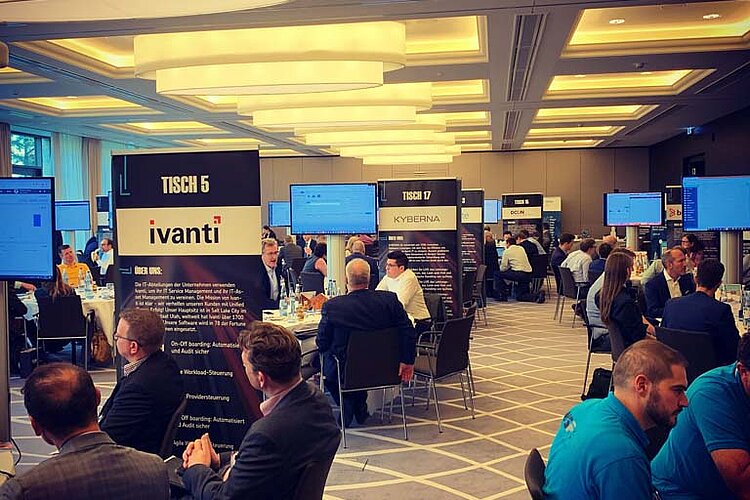 Tool MeetUp participants sit at round tables and discuss with manufacturers while presentations are shown on large screens.