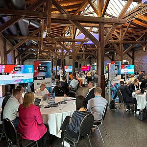 Participants sit at a round table during a tool meetup session, discuss and watch a presentation on a large screen.