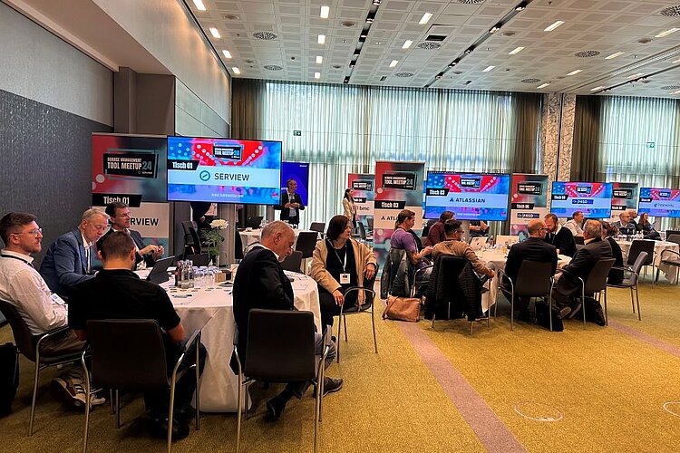 Participants at SERVIEW TOOL MEETUP24 sit at round tables and discuss with manufacturers while presentations are shown on large screens.