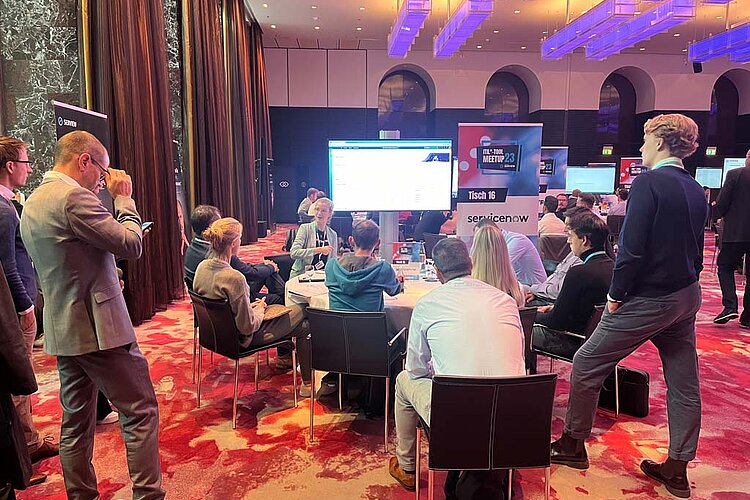 Participants sit at a round table during a tool meetup session, discuss and watch a presentation on a large screen.