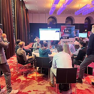 Participants sit at a round table during a tool meetup session, discuss and watch a presentation on a large screen.