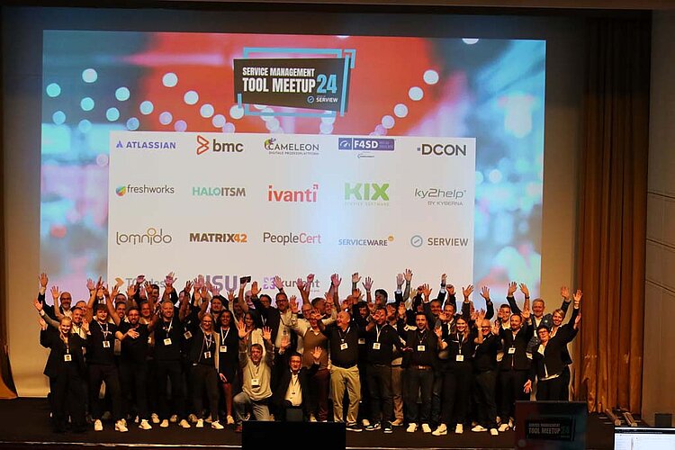 Group photo on the stage of the SERVIEW TOOL MEETUP24 with participants cheering in front of a large screen with sponsor logos.