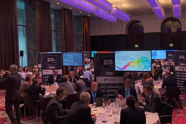 Participants sit at a round table during a tool meetup session, discuss and watch a presentation on a large screen.