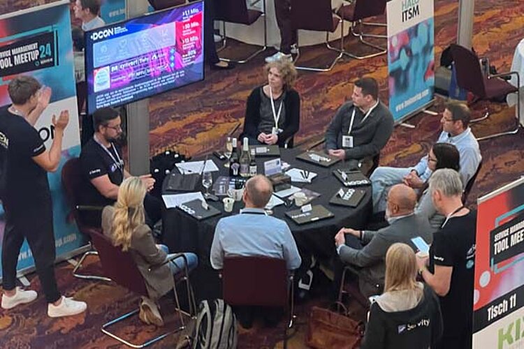 Participants sit at a round table during a tool meetup session, discuss and watch a presentation on a large screen.