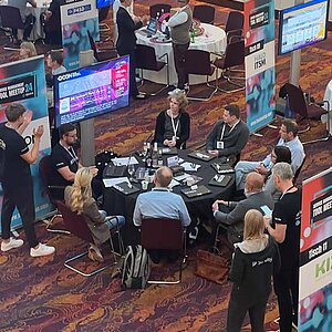 Participants sit at a round table during a tool meetup session, discuss and watch a presentation on a large screen.