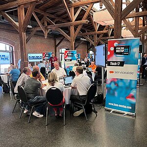 Participants sit at a round table during a tool meetup session, discuss and watch a presentation on a large screen.