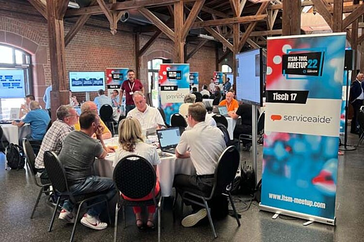 Participants sit at a round table during a tool meetup session, discuss and watch a presentation on a large screen.