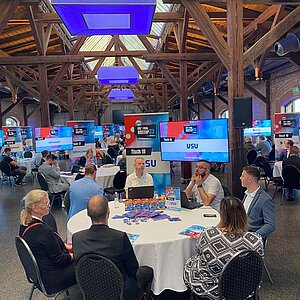 Participants sit at a round table during a tool meetup session, discuss and watch a presentation on a large screen.