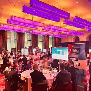 Participants sit at a round table during a tool meetup session, discuss and watch a presentation on a large screen.